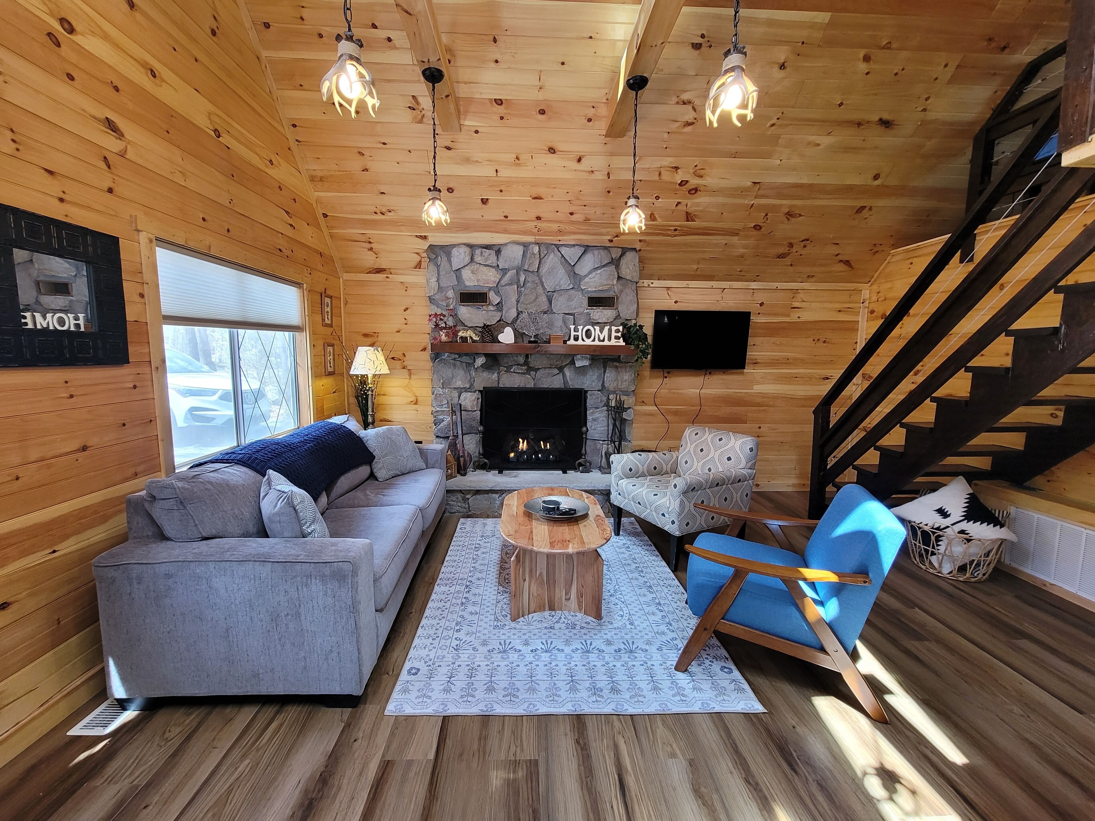 Interior acogedor de una cabaña con chimenea de piedra, paredes de madera, un sofá gris, un tapete estampado y un sillón azul.