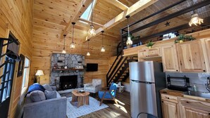Un interior acogedor de cabaña con chimenea de piedra, vigas de madera en el techo, una zona de cocina y un espacio de estar cómodo.