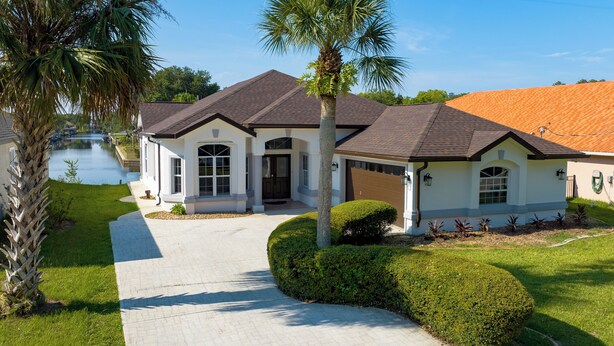 A two-story house with a paved driveway, a palm tree, and a well-maintained lawn. - Five Star 3bd Waterfront Canal Home w/ Private Pool/Spa & Boat Dock. (Palm Coast)