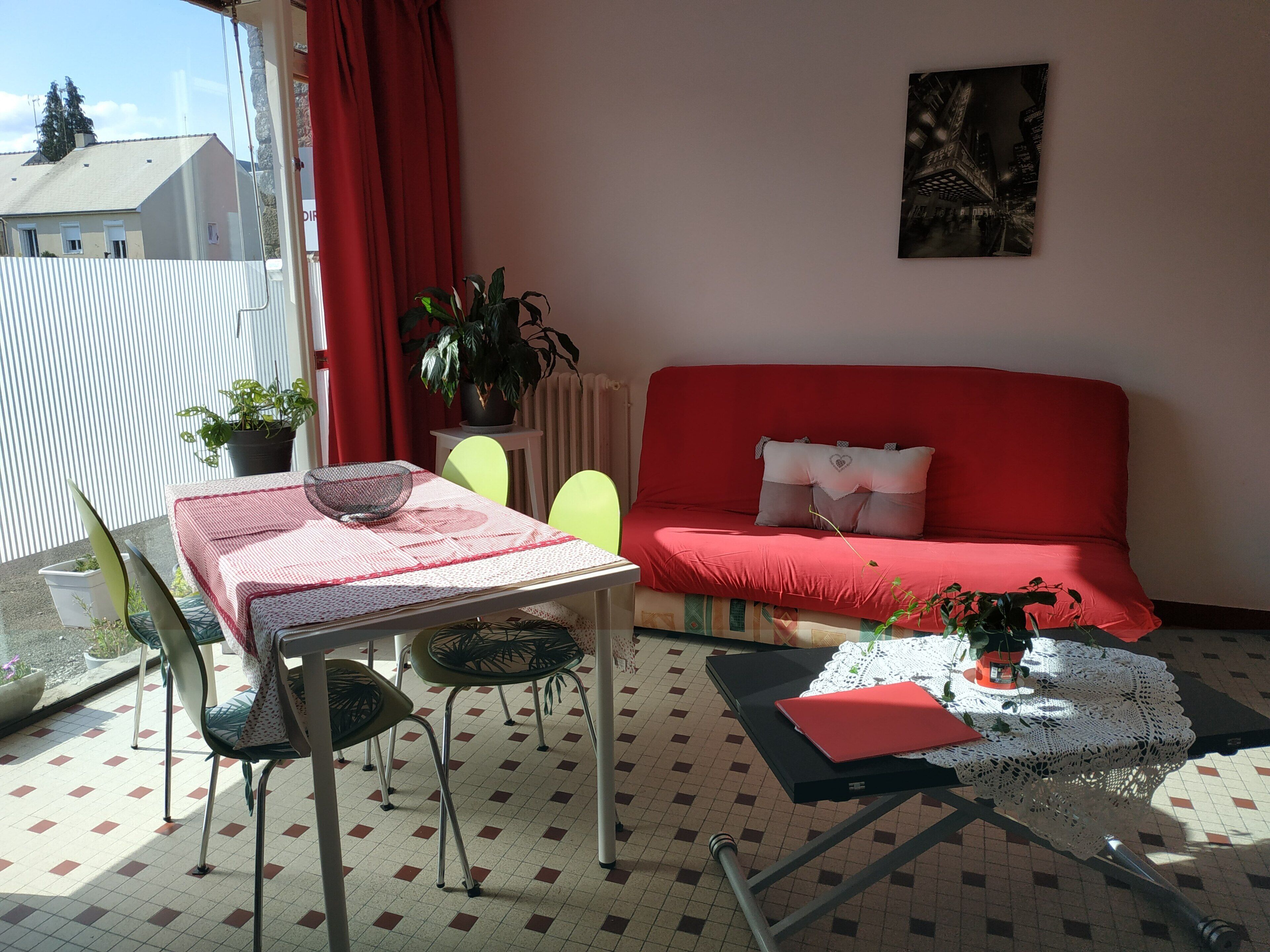 Una sala de estar con un sofá rojo, una mesa de vidrio y un mantel a cuadros rojo y blanco. Hay dos sillones, una planta en maceta y un cuadro en la pared.