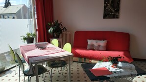 A living room with a red sofa, a glass table, and a red and white checkered tablecloth. There are two chairs, a potted plant, and a framed picture on the wall.