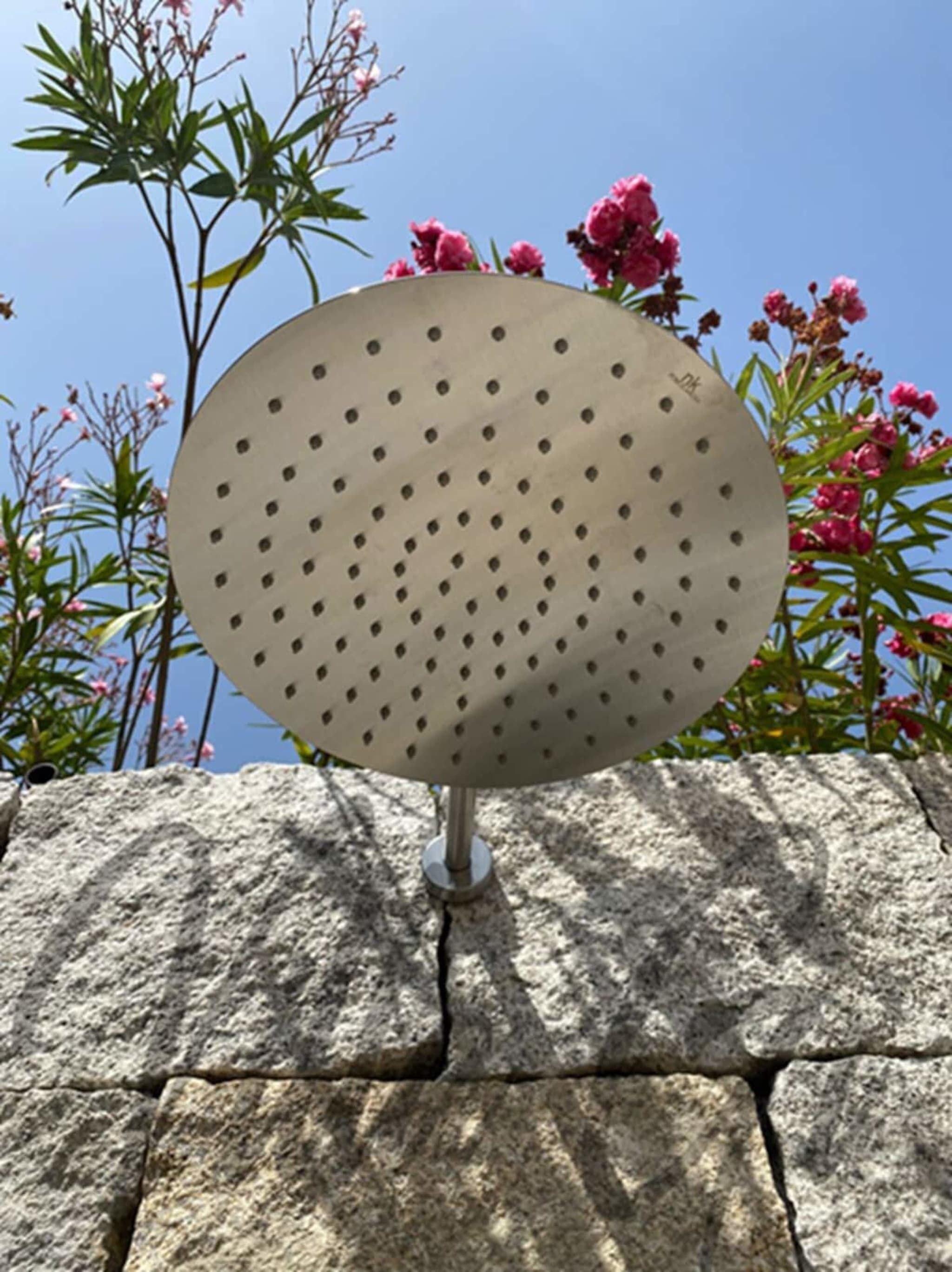 Un objet métallique rond et perforé, fixé sur une surface en pierre, avec un ciel bleu clair et des fleurs roses en arrière-plan.