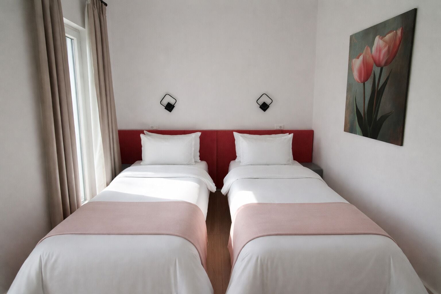 Una habitació d'hotel amb dos llits individuals, un capçal de llit vermell, roba de llit blanca, un quadre de flors i una finestra amb cortines.