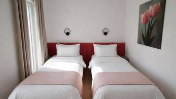 Una habitació d'hotel amb dos llits individuals, un capçal de llit vermell, roba de llit blanca, un quadre de flors i una finestra amb cortines.