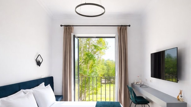 Une chambre d’hôtel moderne, dotée d’un grand lit, d’un bureau, d’une chaise, d’une télévision et d’un balcon offrant une vue sur la verdure.