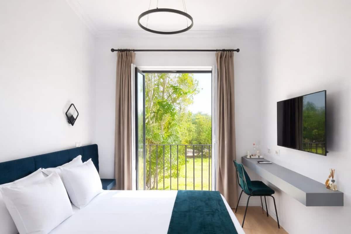 Une chambre d’hôtel moderne, dotée d’un grand lit, d’un bureau, d’une chaise, d’une télévision et d’un balcon offrant une vue sur la verdure.