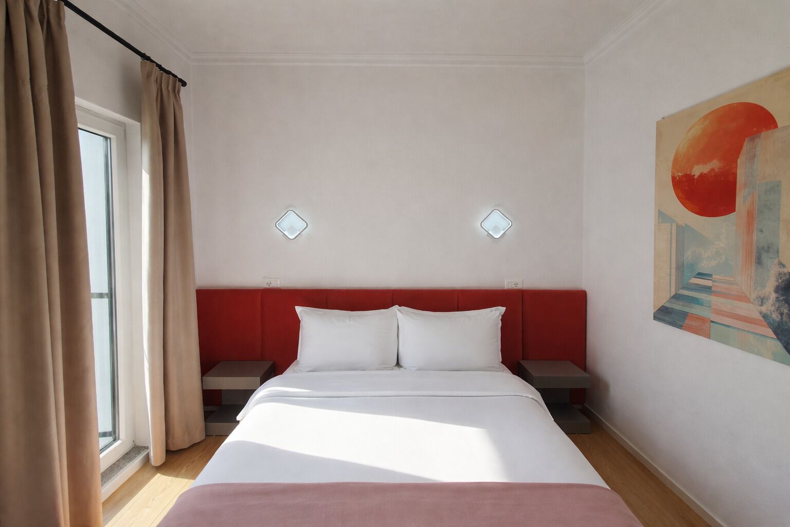 A hotel room with a bed, two bedside tables, a window with curtains, and a wall-mounted light fixture.