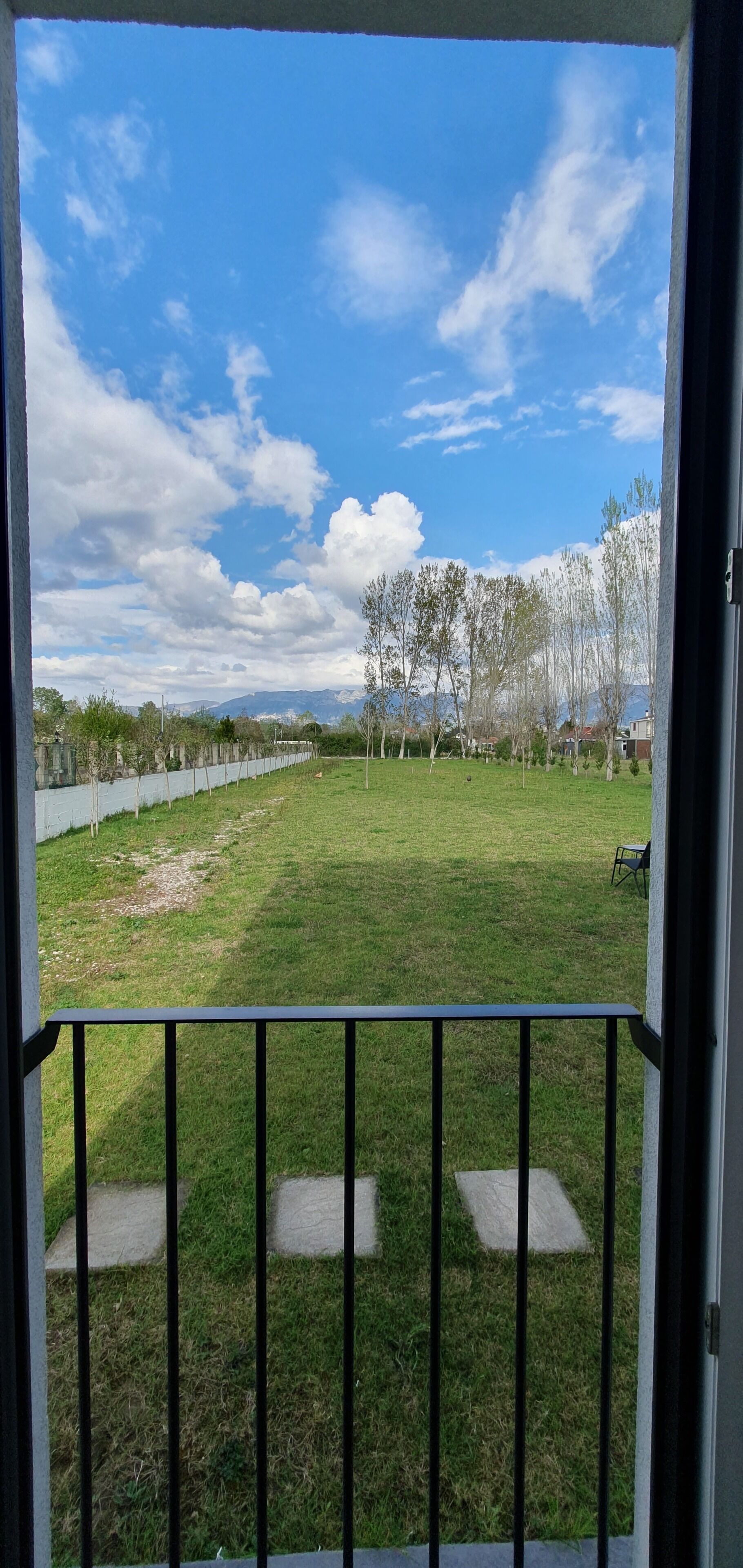 Vista desde un balcón con un jardín cubierto de césped, árboles y cielo despejado.