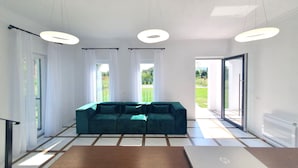 Un salon moderne avec un canapé de couleur turquoise, une table basse en bois et un ordinateur portable posé dessus. On y trouve trois lampes suspendues, des rideaux blancs et une vue sur un jardin, visible à travers la porte vitrée.