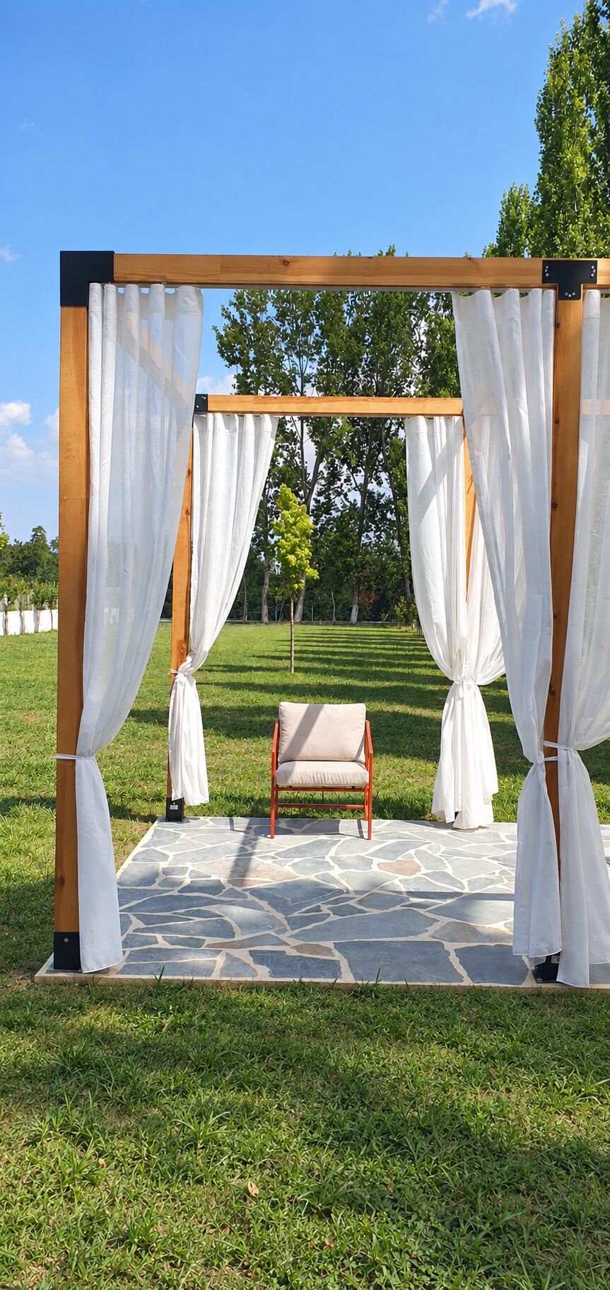 A wooden pergola with white curtains, a stone-tiled floor, and a single chair in a grassy outdoor setting.