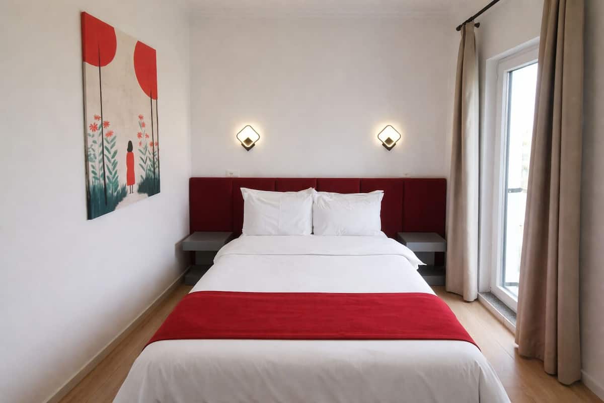 Une chambre à coucher avec un lit, deux tables de chevet, une tête de lit rouge, une fenêtre avec des rideaux et deux lampes fixées au mur.
