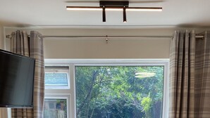 Un coin repas avec une table et des chaises en bois, un téléviseur à écran plat fixé au mur et une vue sur un jardin, visible à travers une grande fenêtre.