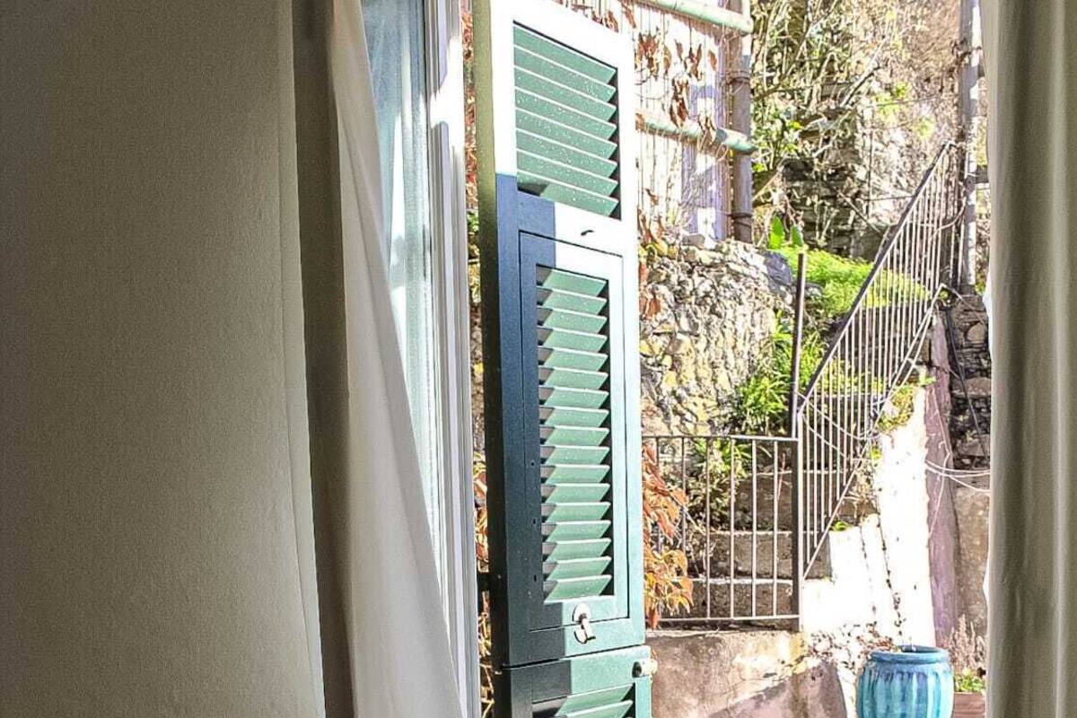 A room with a bed, a wooden table, and a view of a garden through a door with green shutters.