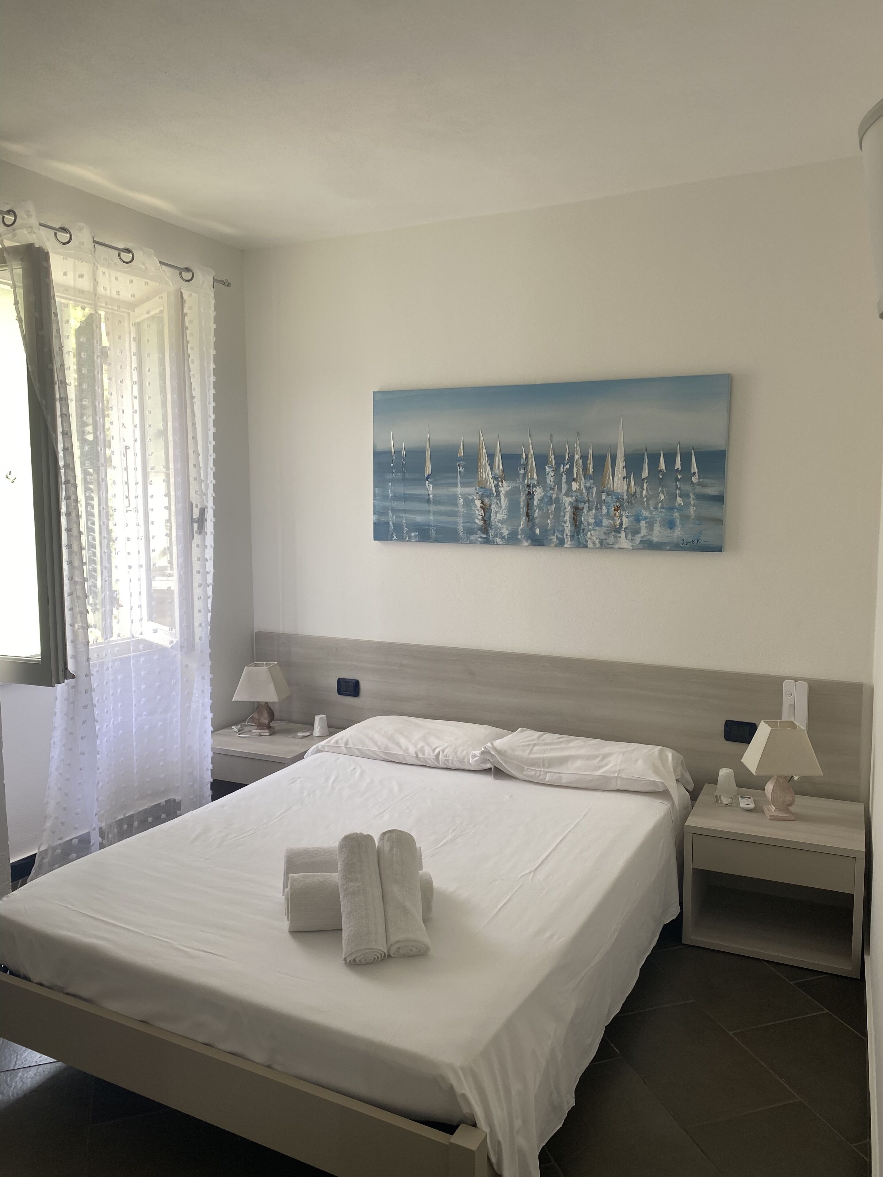 A hotel room with a large bed, two bedside tables, a painting of sailboats, and a window with sheer curtains.