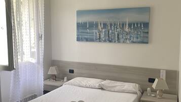 A hotel room with a large bed, two bedside tables, a painting of sailboats, and a window with sheer curtains.