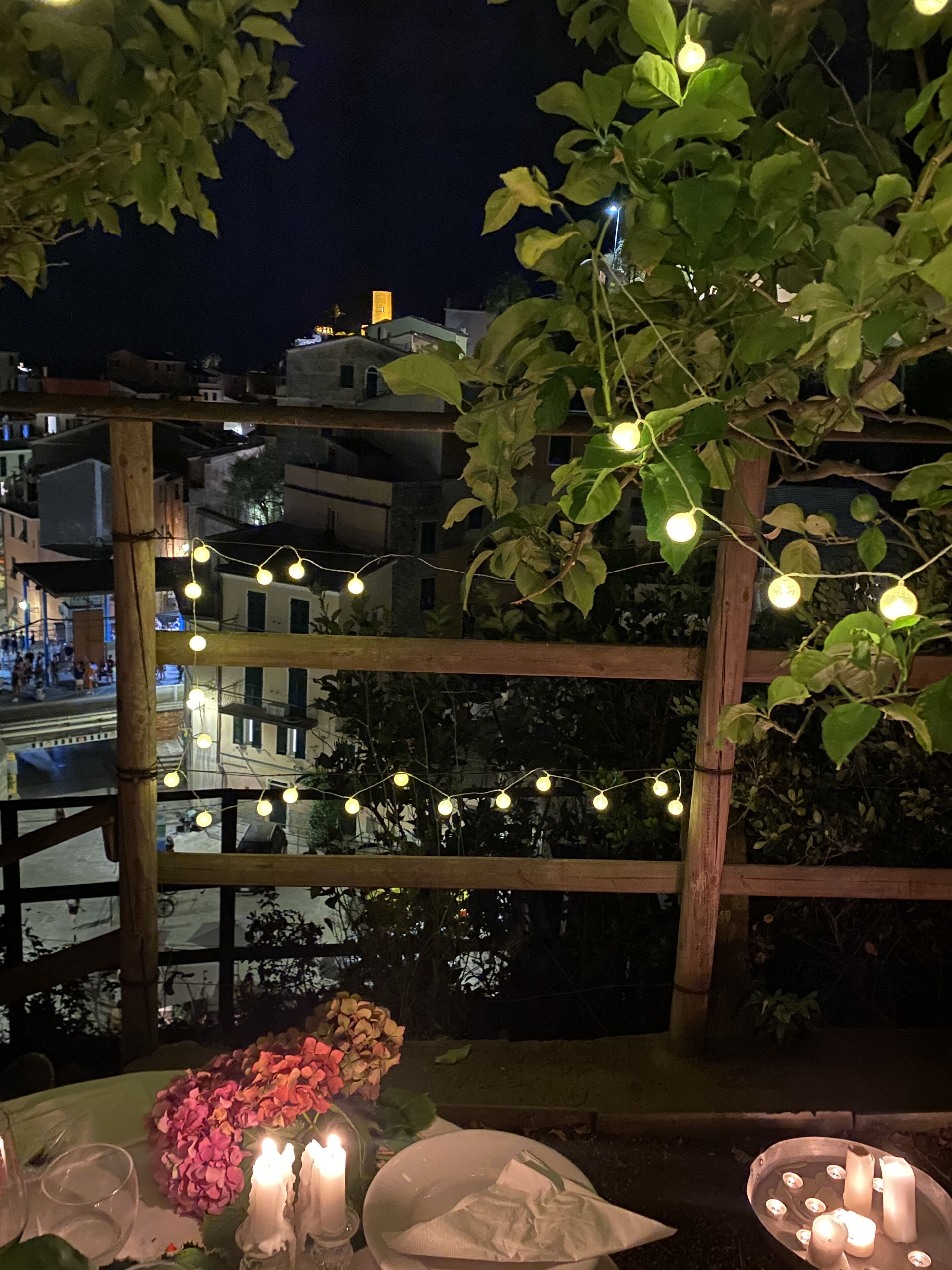 An outdoor dining area with string lights, a table set with candles and flowers, and a view of buildings in the background.
