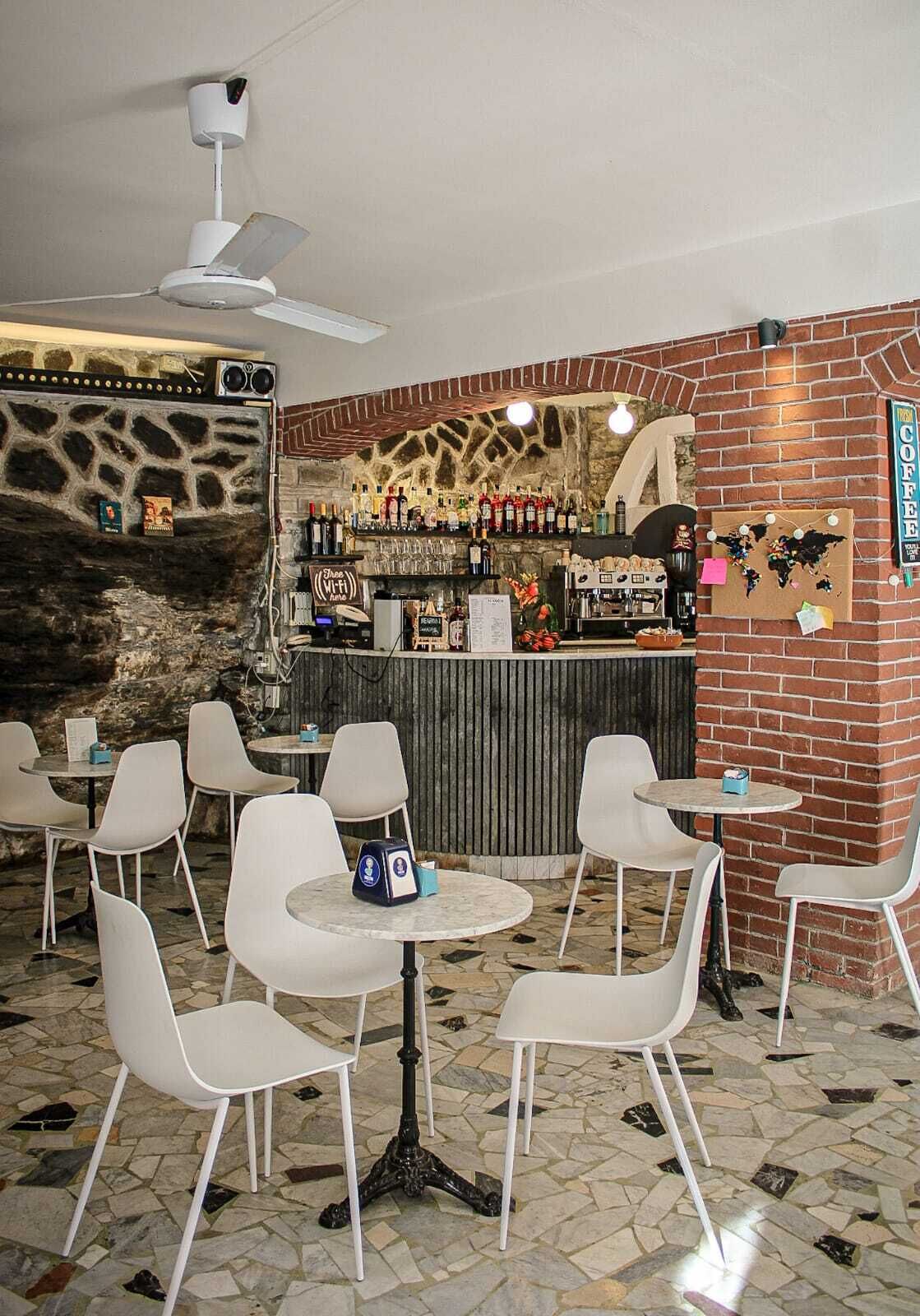 Un restaurante con pared de piedra, pared de ladrillo con un letrero de café, y una zona de bar con varias botellas y vasos. Hay sillas y mesas blancas, y un ventilador de techo.