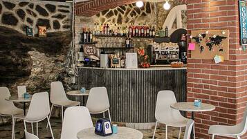 Un restaurante con pared de piedra, pared de ladrillo con un letrero de café, y una zona de bar con varias botellas y vasos. Hay sillas y mesas blancas, y un ventilador de techo.