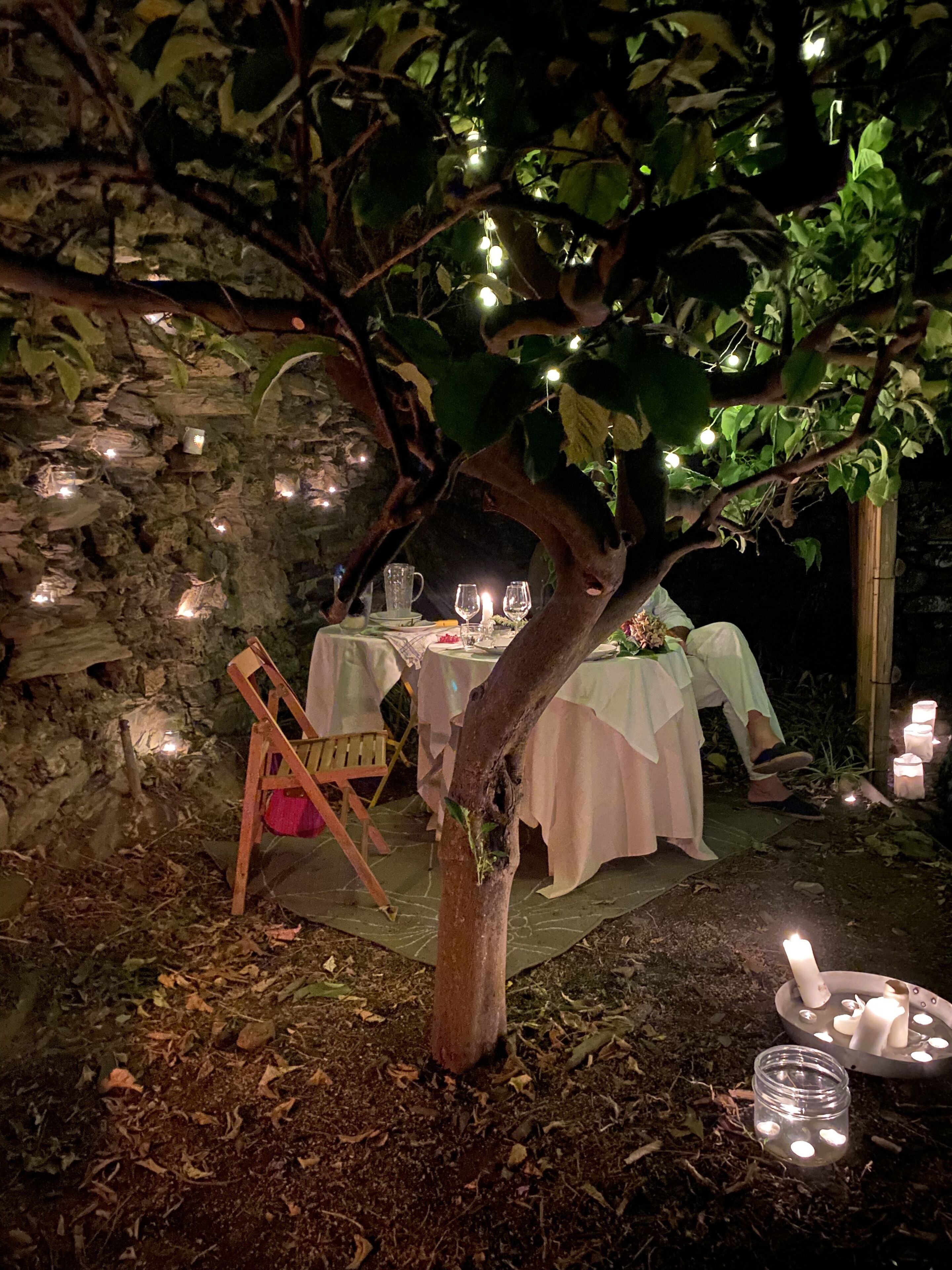 An outdoor dining area with tables set under a tree, surrounded by stone walls and illuminated by string lights.