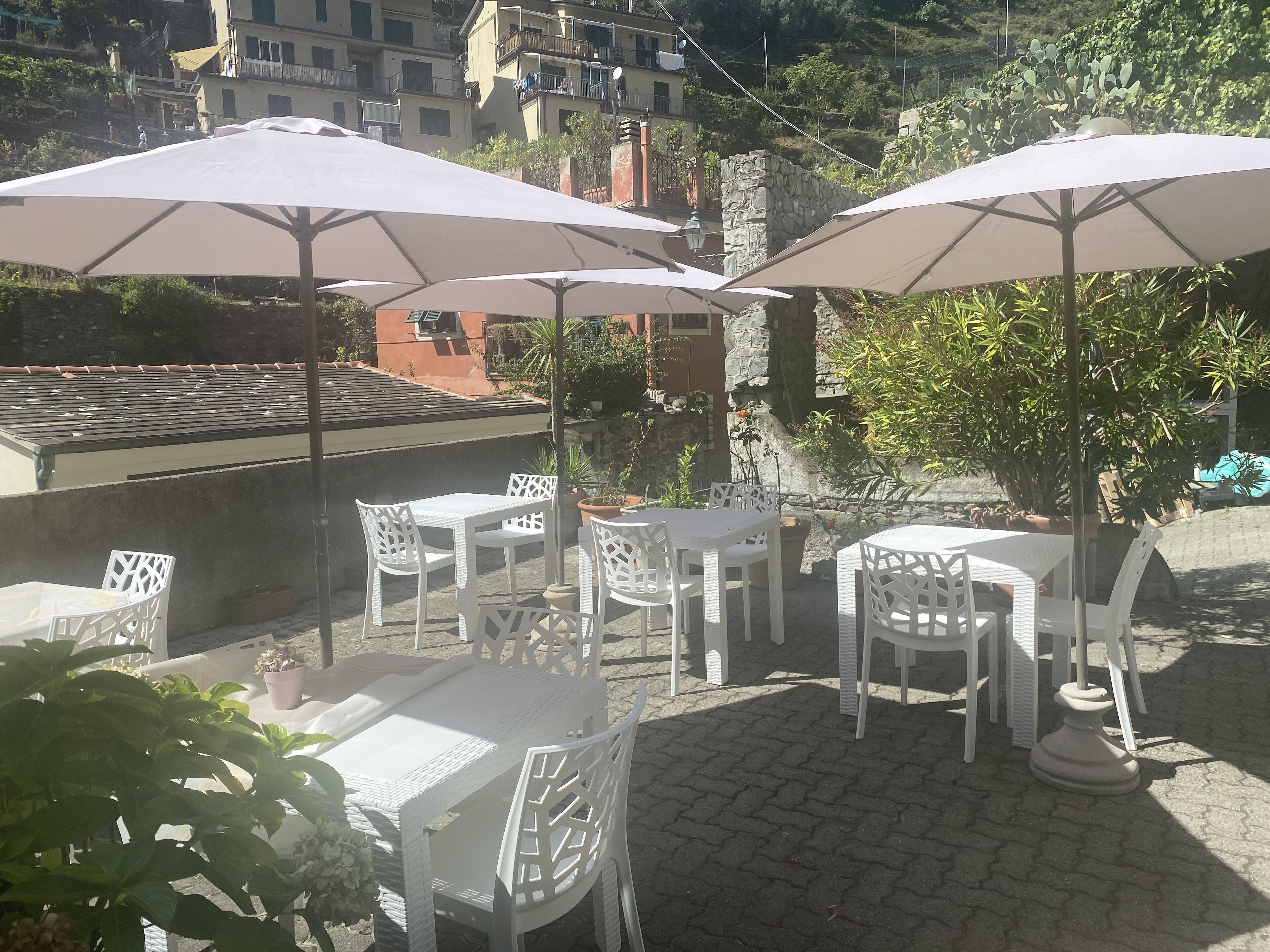 Mobiliario de patio al aire libre con sillas y mesas blancas, cubierto por sombrillas grandes, con edificios y follaje de fondo.
