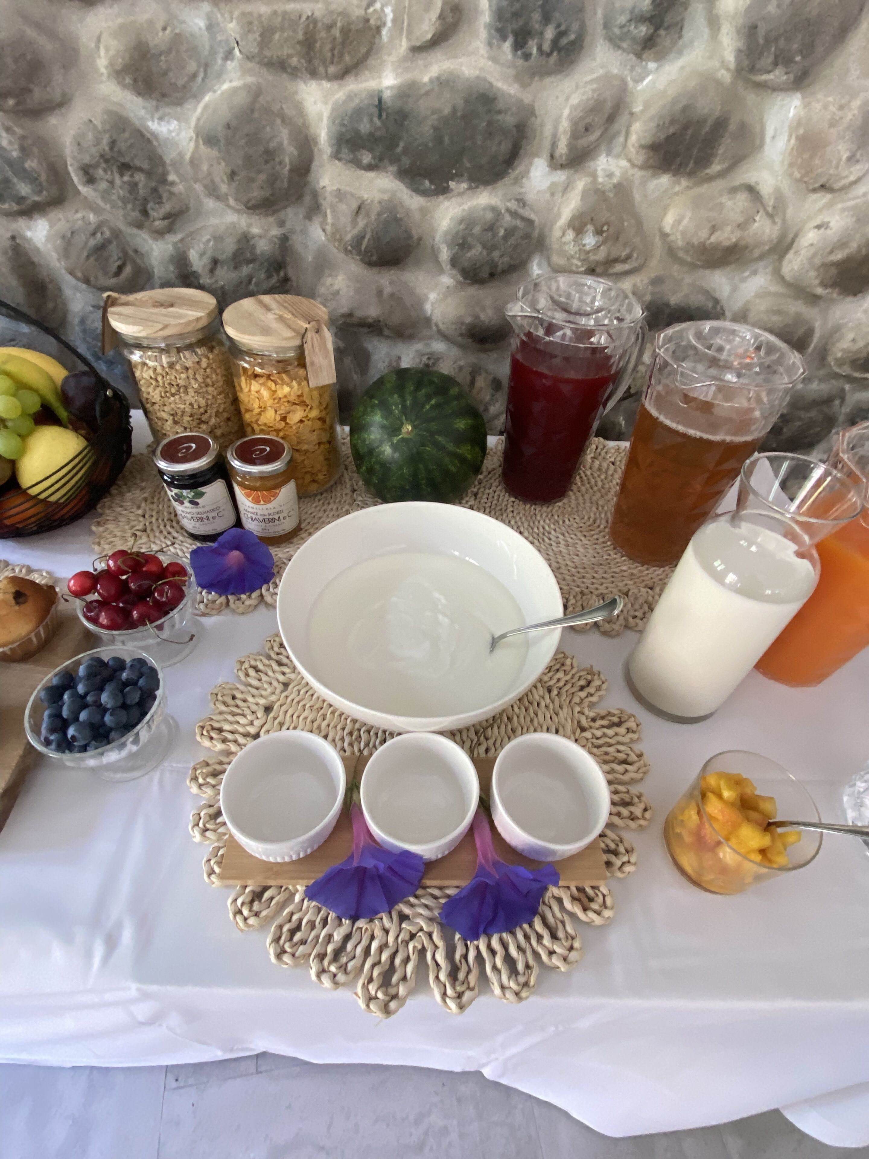 A table with a variety of food items, including fruits, jars of preserves, and drinks, set against a stone wall.