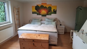 Ein Schlafzimmer mit Bett, Nachttischen, Kommode, Holzkiste, einer Leiter, einem Gemälde und einem Fenster.