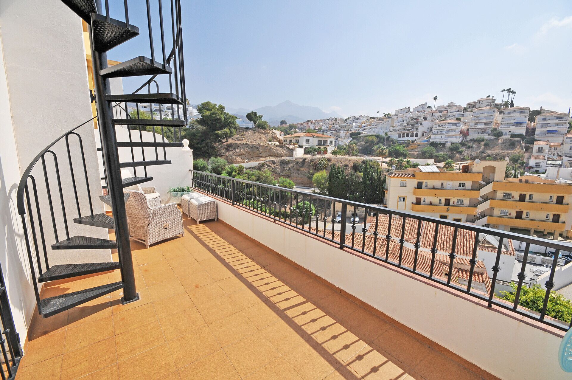 Un balcon doté d’un escalier en métal, d’un sol carrelé et offrant une vue sur une ville côtière, avec ses bâtiments et une montagne en arrière-plan.