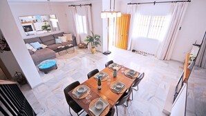 A dining area with a wooden table set for four, surrounded by chairs. There is a sofa and a small round table with a blue cushion in the background. - Pinomar 1A (Nerja)