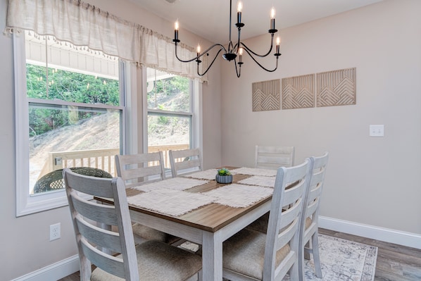 A dining area with a wooden table, white chairs, a chandelier, and a view of a garden through large windows.