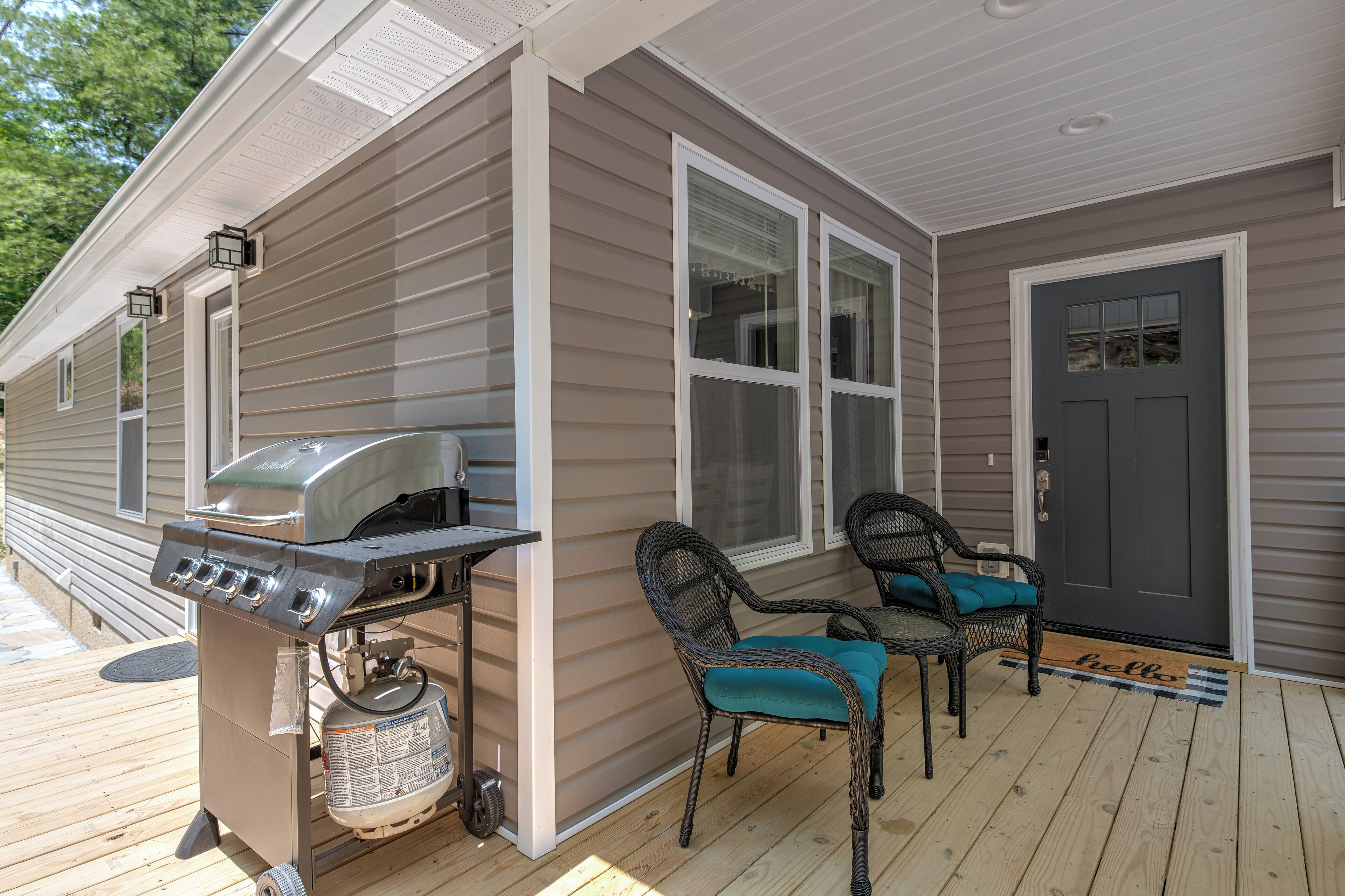 Un porche abrité avec un barbecue, deux fauteuils en osier et une porte sur laquelle est posé un paillasson avec l'inscription « Bonjour ».