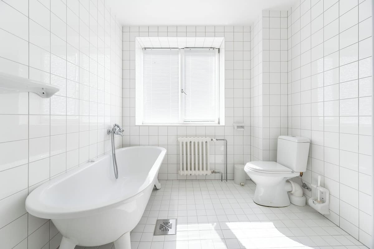 A modern bathroom with white tiles, a freestanding bathtub, a toilet, and a window with blinds.