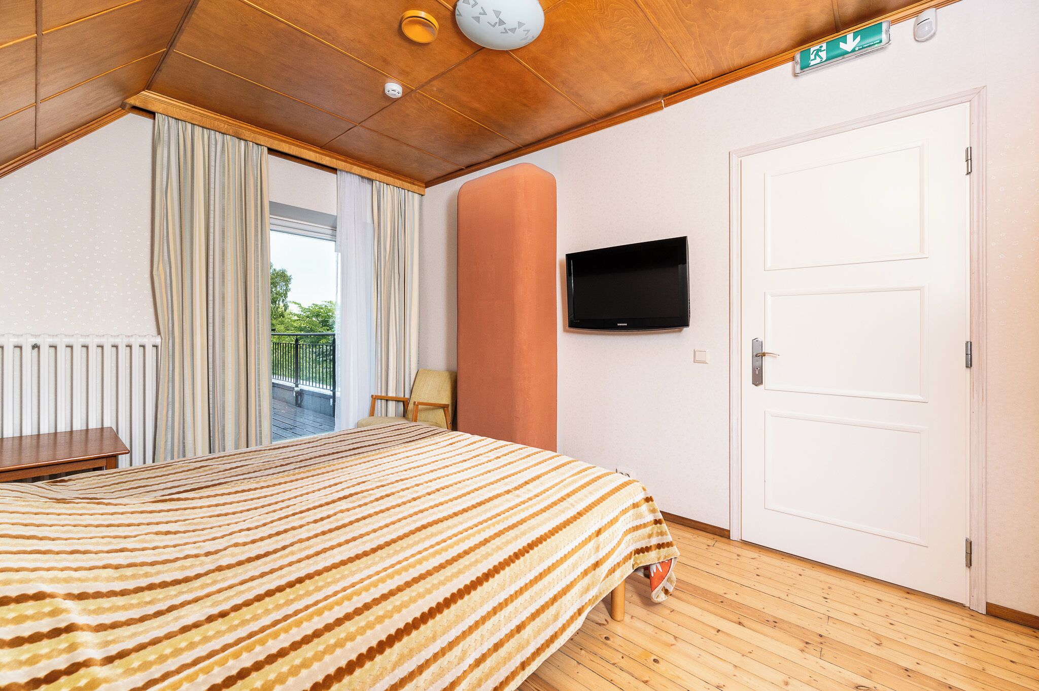 A hotel room with a bed, a television mounted on the wall, a door, and a view of the outdoors.