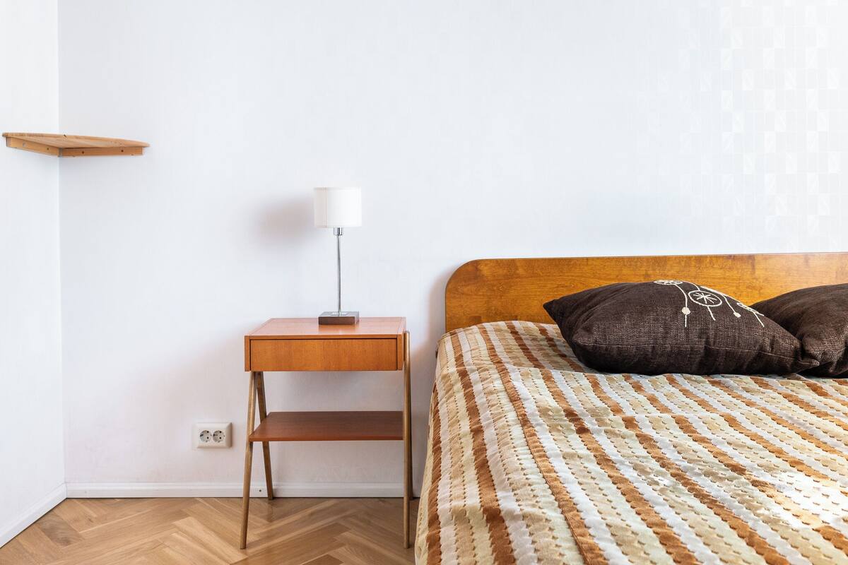 A bedroom with a wooden headboard, a bed with striped bedding, a bedside table with a lamp, and a small shelf.