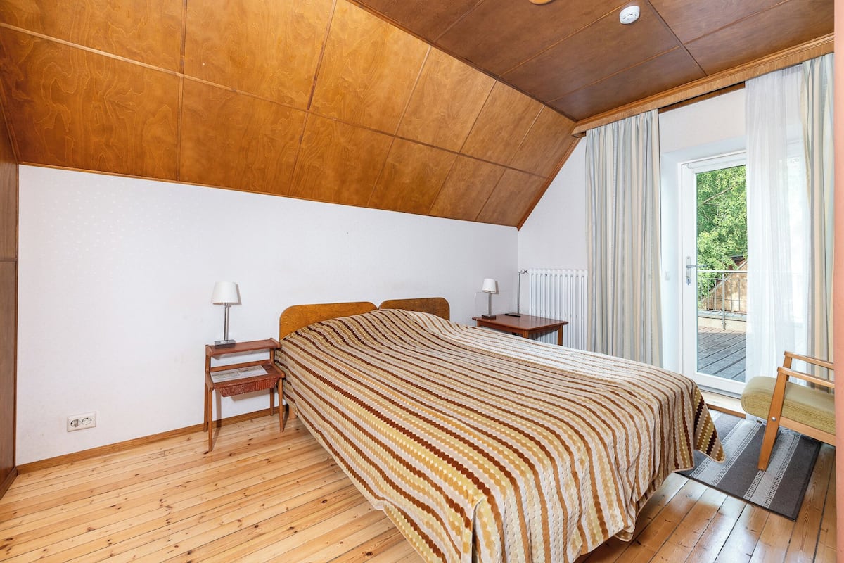 A bedroom with a wooden ceiling, a large bed with a striped bedspread, a nightstand with a lamp, a chair, and a view of the outdoors through a window.