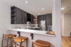 A modern kitchen with dark cabinets, a white countertop, and stainless steel appliances. There are three wooden bar stools with cushioned seats. - Le Chamonix A 2 Bedroom Condo (Mountain Village)