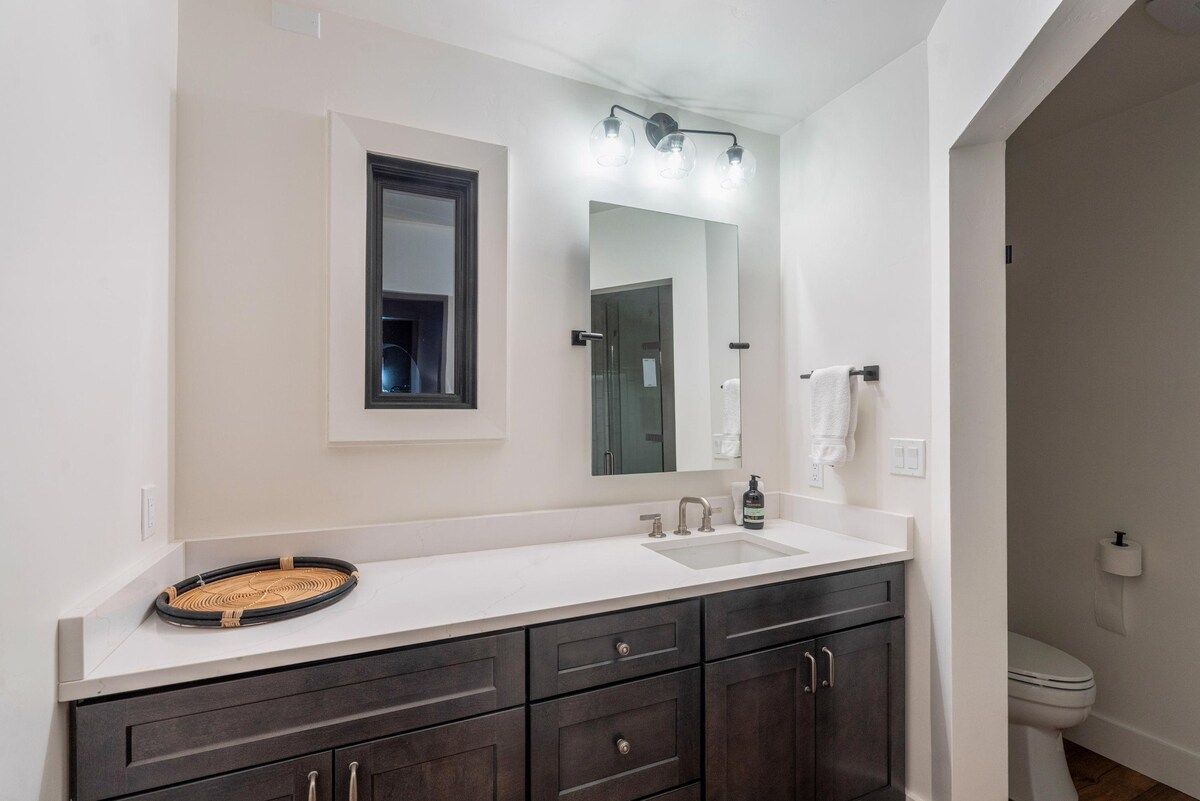 A modern bathroom with a white vanity, dark cabinets, a large mirror, and a toilet.