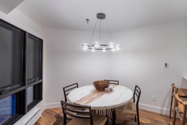 A dining area with a round table, chairs, a central light fixture, and a window with blinds. - Le Chamonix A 2 Bedroom Condo (Mountain Village)