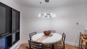 Et spiseområde med et rundt bord, stole, en centralt placeret lampe og et vindue med persienner.