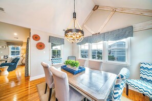 A dining area with a wooden table, two chairs, a chandelier, and a window with patterned curtains. - Spacious 5-Bed, 3-Bath Corner Condo, Steps to Beach, Boardwalk & Downtown! (Ocean City)