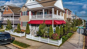 A two-story house with a red awning, white picket fence, and a well-maintained front yard with trimmed bushes and a small tree. - Spacious 5-Bed, 3-Bath Corner Condo, Steps to Beach, Boardwalk & Downtown! (Ocean City)
