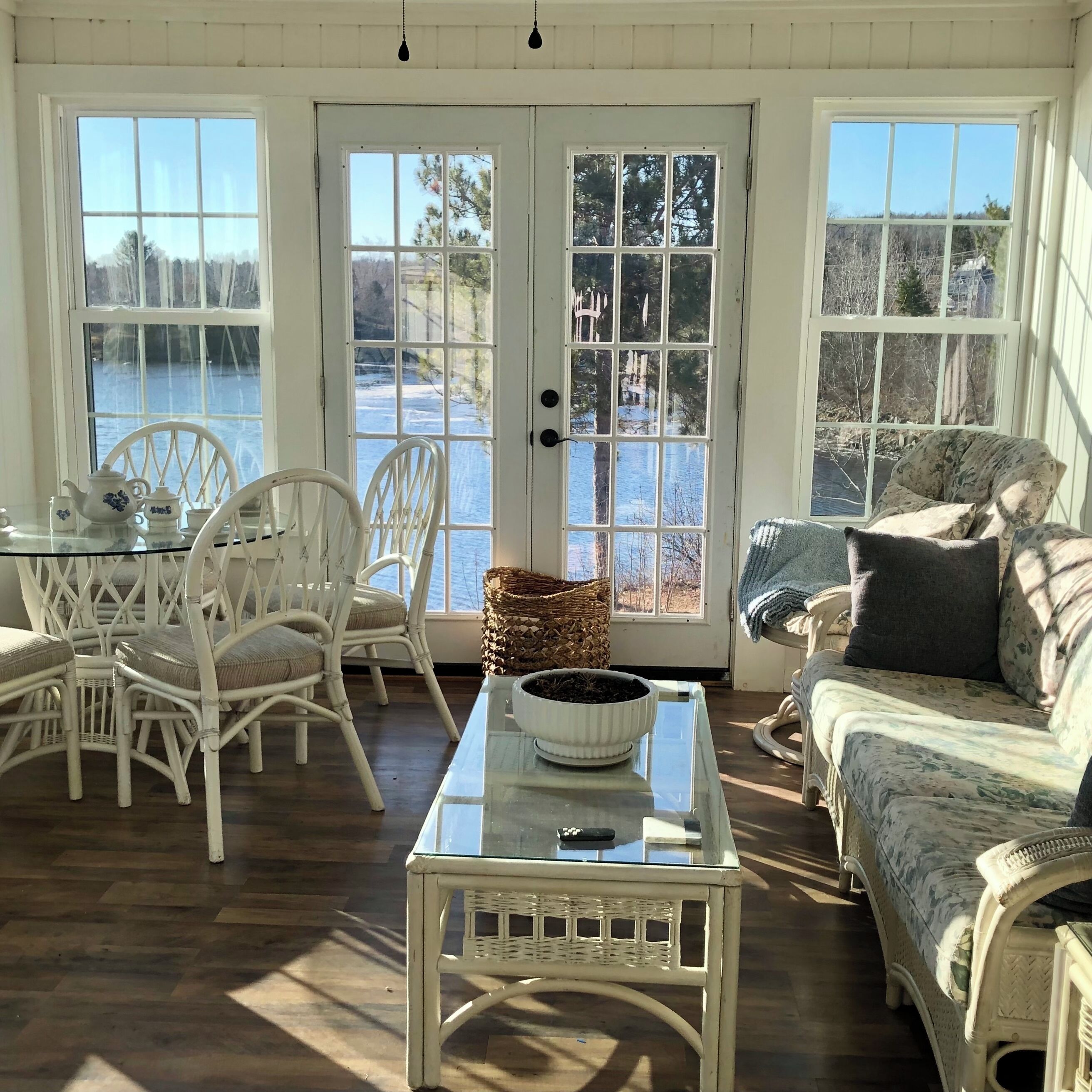 A sunlit room with a table, chairs, a sofa, and a view of a body of water through large windows.