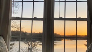Huone, jossa on suuri ikkuna, josta avautuu näkymä vesistöön auringonlaskun aikaan, sänky tyynyineen ja peitto.