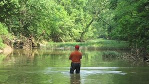 Un hombre de pie en un río rodeado de abundante vegetación.
