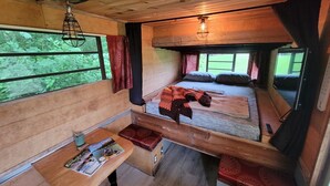 Una cabaña acogedora con interior de madera, una cama con manta, una mesa con una revista y una ventana con vistas a la vegetación.
