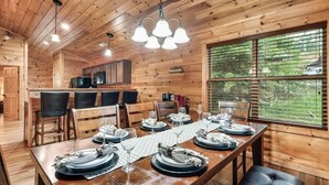A wooden cabin interior with a dining area set for four, a kitchen, and large windows. - Free Dollywood Tix/Pool/5Min2Dollywood/Pool Table (PIgeon Forge)