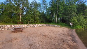 A sandy beach with a wooden picnic table and a stone wall, surrounded by dense forest and a calm body of water. - Northwoods Lodge Hotel Room 1 (Long Lake)