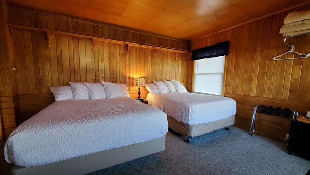 A wooden-paneled room with two beds, a window with blinds, a hanging clothes rack, and a radiator. - Northwoods Lodge Hotel Room 1 (Long Lake)