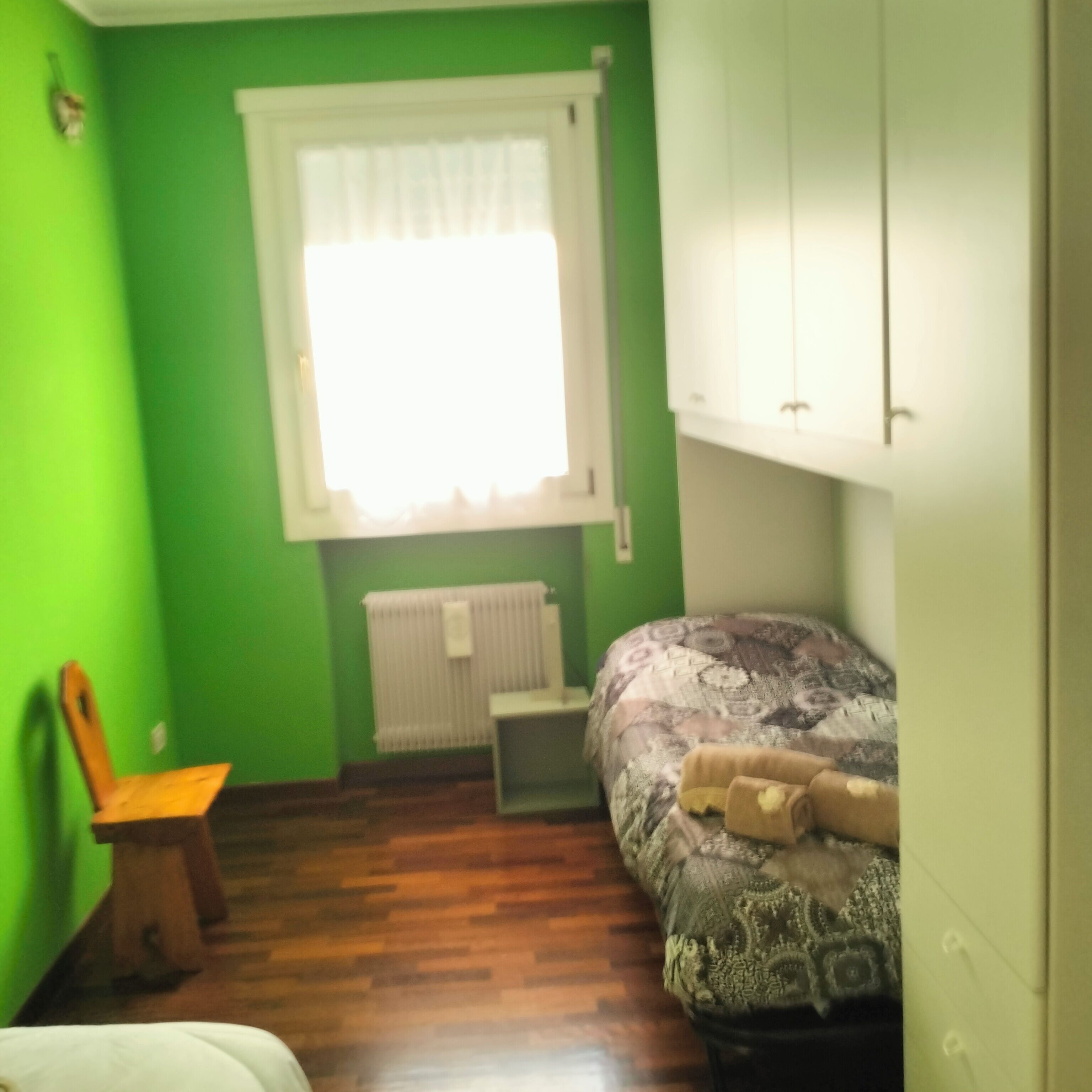 A small bedroom with green walls, a window with white curtains, a radiator, a bed with patterned bedding, and a wooden chair.