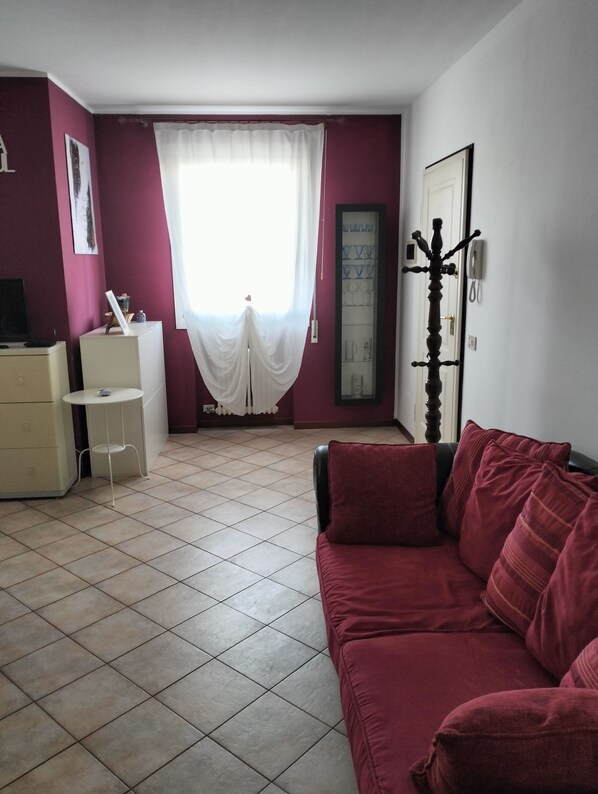 Una habitación con un sofá rojo, una mesa blanca y un armario. Hay una ventana con cortinas blancas y una puerta con perchero.