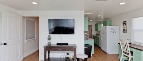 Una cocina moderna con un televisor de pantalla plana sobre un mueble bajo de madera, un refrigerador y una zona de comedor con una silla verde.